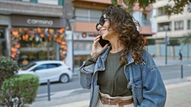 Kıvırcık saçlı, güneş gözlüğü takan ve kot ceket giyen gülümseyen İspanyol kadın şehir merkezinde telefonla konuşuyor.