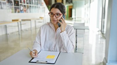 Beyaz tişörtlü, odaklanmış bir İspanyol kadın içeride belgelerle çalışırken telefonla konuşuyor..