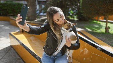 Genç bir kadın güneşli bir parkta selfie çekerken köpeğini sevgiyle öpüyor..