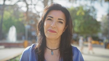 Olgun İspanyol kadın güneşli bir parkta güvenle poz veriyor. Bulanık yeşillik ve çeşme arkaplanı var..