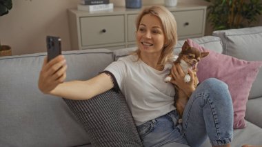 Günlük giysiler içinde genç sarışın beyaz bir kadın oturma odasında oturmuş Chihuahua evcil hayvanıyla selfie çekiyor..