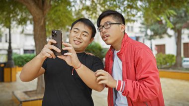 Arka planda bulanık yeşillik olan bir parkta akıllı telefon kullanan iki Asyalı adam..