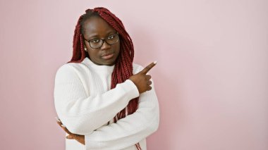 Saçları örgülü, pembe arka planda beyaz süveter giyen, kendine güvenen Afro-Amerikan kadın.