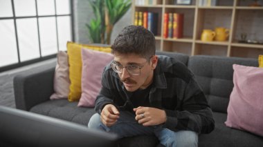 Gözlüklü ve bıyıklı genç bir İspanyol adam rahat bir oturma odasında dikkatle televizyon izliyor..