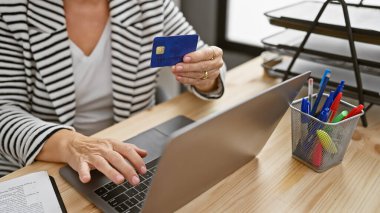 Ofis masasında dizüstü bilgisayar kullanan orta yaşlı bir kadın kredi kartı tutuyor..