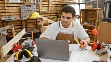 A focused young man with a beard in a carpentry workshop using a laptop and measuring tape. clipart