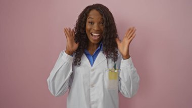 Güzel, genç, kıvırcık saçlı ve laboratuvar önlüklü Afro-Amerikan bir kadın, izole edilmiş pembe bir arka plan için heyecanını dile getiriyor, neşe ve profesyonelliği somutlaştırıyor..