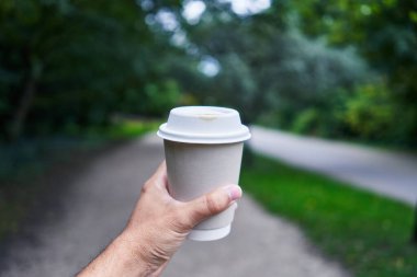 Yeşil bir parkta yürürken tek kullanımlık kahve fincanı tutan bir adam.