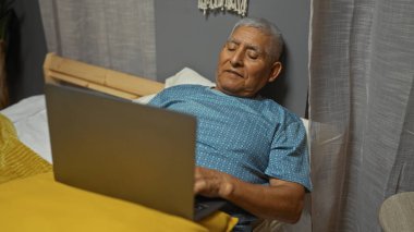 Beyaz saçlı İspanyol bir adam yatak odası ortamında dizüstü bilgisayarda çalışıyor, rahat bir ev ortamında yatakta uzanıyor, olgun, rahat bir ev hayatı yaşıyor..