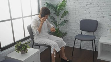 Modern bir kapalı bekleme odasında oturan, yaşlı, kısa saçlı, İspanyol bir kadın bir telefonla saatini ciddi bir ifadeyle kontrol ediyor..