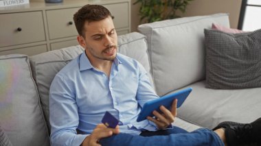 Sakallı, yakışıklı, genç, İspanyol bir adam evinin oturma odasında elinde bir kredi kartı ve mavi bir tabletle açık mavi bir gömlek giyerken oturuyor..