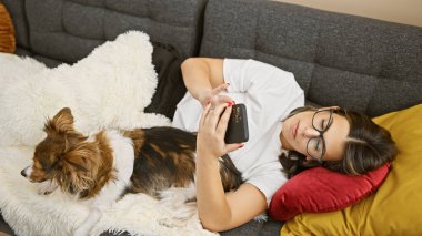 Evde kanepede köpekle kucaklaşırken akıllı telefon kullanan rahat bir kadın.