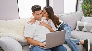 Bir kadın, erkeğin yanağından öpüyor. Bir kanepede dizüstü bilgisayarla dinlenirken, modern aşkı ve ev rahatlığını yansıtıyor..