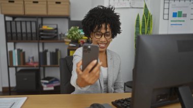 Kıvırcık saçlı, güzel, Afro-Amerikan bir kadın iş günü modern ofis ortamında selfie çekerken gülümsüyor..