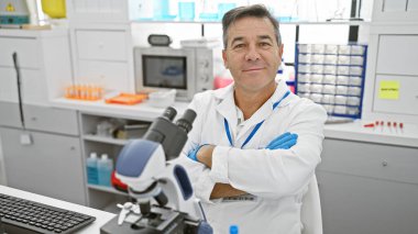 Laboratuvar önlüklü, kendinden emin orta yaşlı bir adam modern bir laboratuvar ortamında kollarını kavuşturdu..