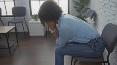 Kıvırcık saçlı güzel, Afro-Amerikalı bir kadın kapalı bir bekleme odasında endişeli endişeli oturuyor..