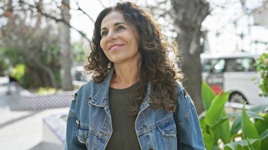 Kıvırcık saçlı, gülümseyen, olgun bir İspanyol kadın güneşli bir parkta doğanın tadını çıkarıyor..