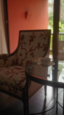 Cam masanın yanında dekoratif desenli zarif bir sandalye. Kapalı alanda kahve fincanı ve doğal ışıklandırma..