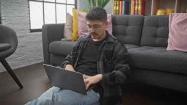 Bıyıklı genç bir İspanyol adam evinde dizüstü bilgisayarda çalışıyor. Yerlerde, dekore edilmiş oturma odasındaki kanepenin karşısında rahatça oturuyor..