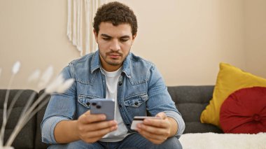 Sakallı genç adam rahat bir oturma odasında online alışveriş için akıllı telefon ve kredi kartı kullanıyor..