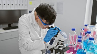 Eldiven giyen bir erkek bilim adamı, iyi donanımlı bir laboratuvarda mikroskopla bir örneği inceliyor..