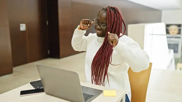 stock image Smiling black woman with braids wearing glasses at her workplace, celebrating success in front of a laptop.