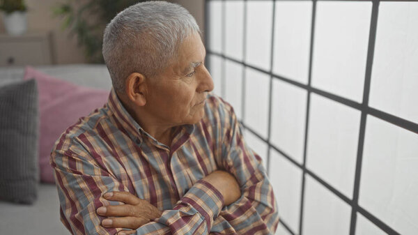 Mature grey-haired hispanic man with crossed arms looking thoughtfully out a window in a cozy living room.