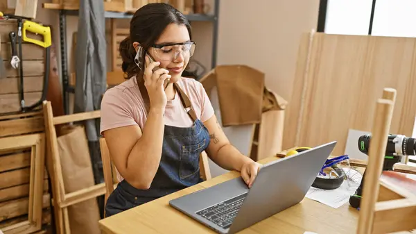 Stock image Hispanic woman in a carpentry workshop communicating on phone while using laptop.