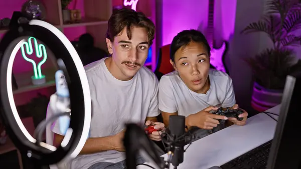 stock image Interracial couple gaming together in a dark room with neon lights and streaming equipment, focusing intensely on the screen.