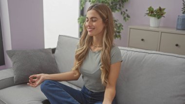 Rahat, genç bir kadın, sarışın, gülümseyen ve meditasyon yapan modern bir oturma odasında.