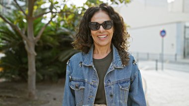 Kıvırcık saçlı, güneş gözlüğü takan ve şehir parkında kot ceket giyen gülümseyen orta yaşlı bir kadın..