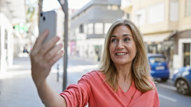 Gülümseyen orta yaşlı beyaz kadın şehir sokaklarında selfie çekiyor.