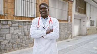 Tıbbi ceket ve steteskop giymiş kendine güvenen Afrikalı bir adam şehir caddesinde kollarını kavuşturmuş duruyor..