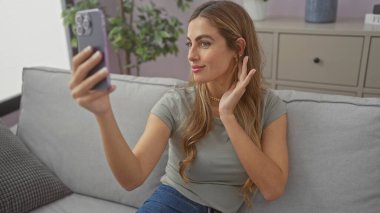 Kafkasyalı genç bir kadın evinde akıllı telefonuyla selfie çekiyor. Günlük kıyafetleriyle gülümsüyor..