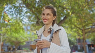 Yeşil bir park şehrinde telefon kullanan gülümseyen beyaz bir kadın..