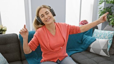 Neşeli orta yaşlı bir kadın kulaklıkla müzik dinlerken evde dans eder, mutluluk ve rahatlığı somutlaştırır..
