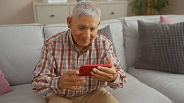 Yakışıklı, gri saçlı, İspanyol bir adam rahat oturma odasında rahat bir kanepede otururken akıllı telefonuyla bir video görüşmesinden zevk alıyor..