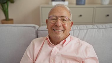 Oturma odasında gülümseyen yaşlı beyaz bir adam, bir kanepede oturuyor, ev dekorunda mutlu ve yakışıklı bir beyefendiyi sergiliyor..