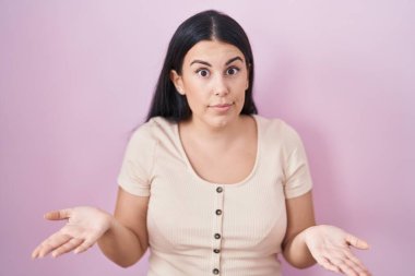 Pembe arka planda duran Latin kökenli genç bir kadın. Kolları açık ve kafası karışık, hiçbir fikri yok.. 