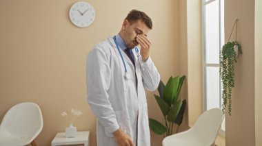 Steteskoplu, beyaz önlüklü yorgun bir erkek doktor hastane bekleme odasında gözlerini ovalıyor. Beyaz sandalyeler, bir saat ve kapalı salonda bitkiler..