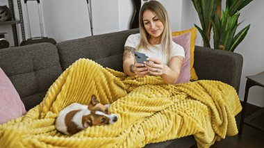 Rahat bir kadın, rahat bir ev ortamında sarı bir battaniyeye uzanmış köpeğiyle akıllı telefon kullanıyor..