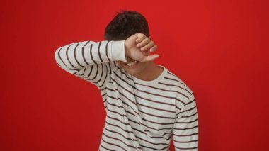 Çizgili tişört giyen yakışıklı bir İspanyol adam yüzünü kırmızı bir duvarın arkasına gizlerken gülümsüyor..