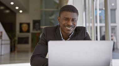 Modern bir ofis içinde dizüstü bilgisayarla çalışırken gülümseyen Afrikalı bir adam profesyonelliği ve iş memnuniyetini resmediyor..