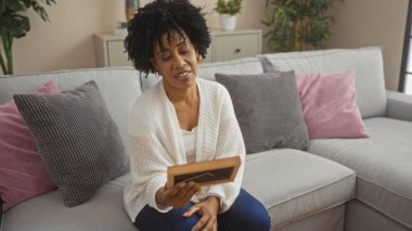 Kıvırcık saçlı güzel bir Afro-Amerikalı kadın, elinde çerçeveli bir fotoğraf tutarak oturma odasında oturuyor. Dolgun yastıklar ve ev bitkileriyle çevrili. Huzurlu bir ev dekorasyonu sergiliyor..