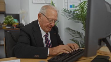 Ofis ortamında klavyeye yazı yazan yaşlı, beyaz bir adam. Kapalı mekanları ve profesyonel kıyafetleri var. Olgun bir iş ortamının altını çiziyor..