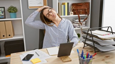 Modern ofis ortamında dizüstü bilgisayar ve evraklarla boynunu uzatan esmer bir kadın..