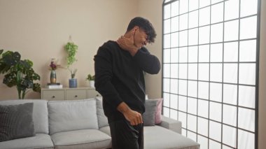 Boynu ağrıyan genç bir İspanyol adam, büyük bir pencereyle karakterize edilen bitki ve modern mobilyalarla dolu şık dekore edilmiş bir oturma odasında duruyor..