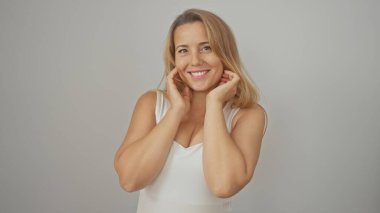 Sarı saçlı, gülümseyen, beyaz bluzlu, gri bir arka plana karşı poz veren beyaz bir kadın.