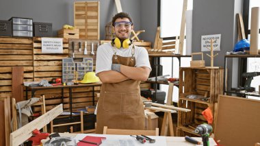 Güvenlik gözlüğü ve kulaklık takan sakallı, gülümseyen genç bir adam bir marangoz atölyesinde kollarını kavuşturuyor..