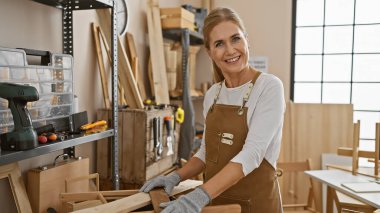 Güneşli bir atölyede ağaçla çalışan önlüklü, gülümseyen kadın marangoz.